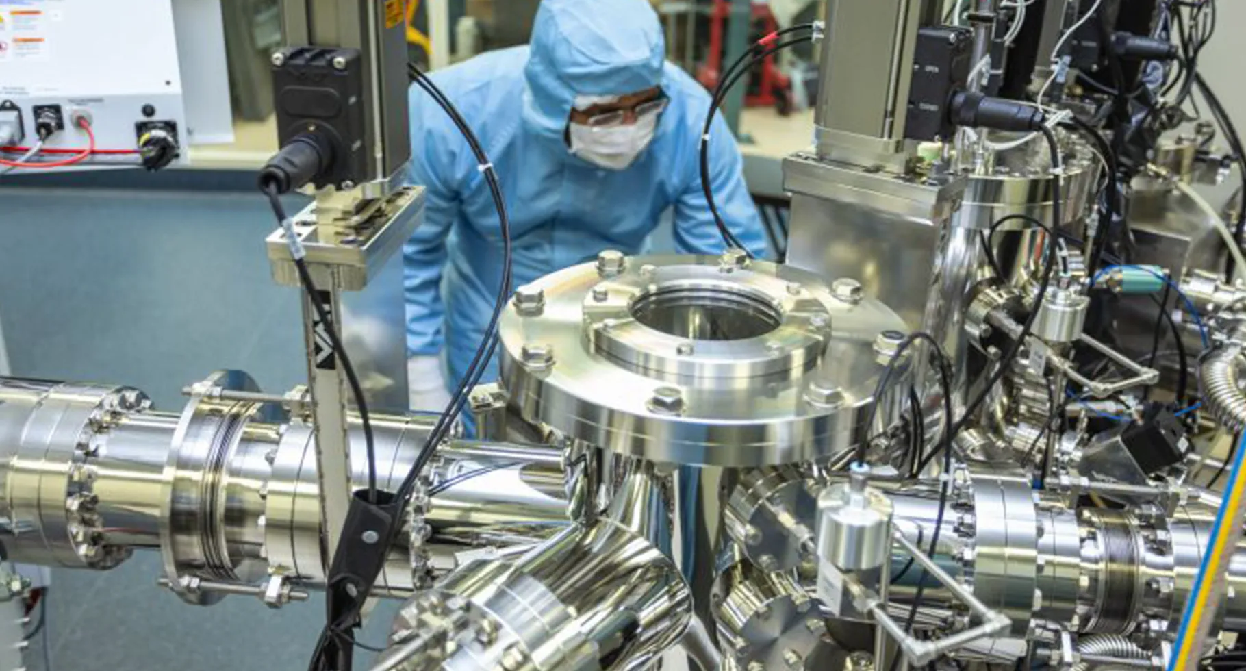 A person wearing a blue clean suit and mask bends over a large metal array of tubing.