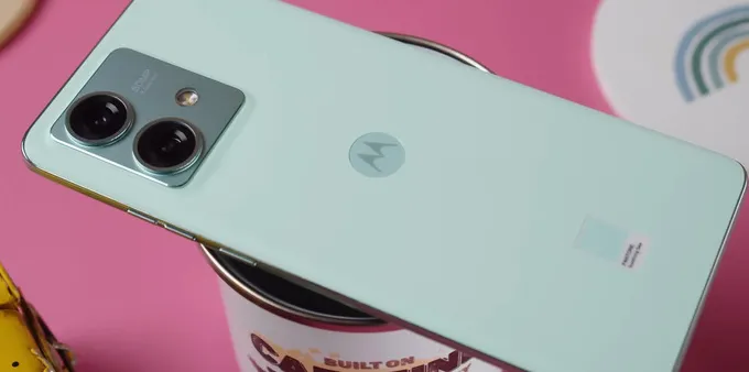 A light green Motorola smartphone sits on top of a coffee cup, both of which sit on a pink backdrop