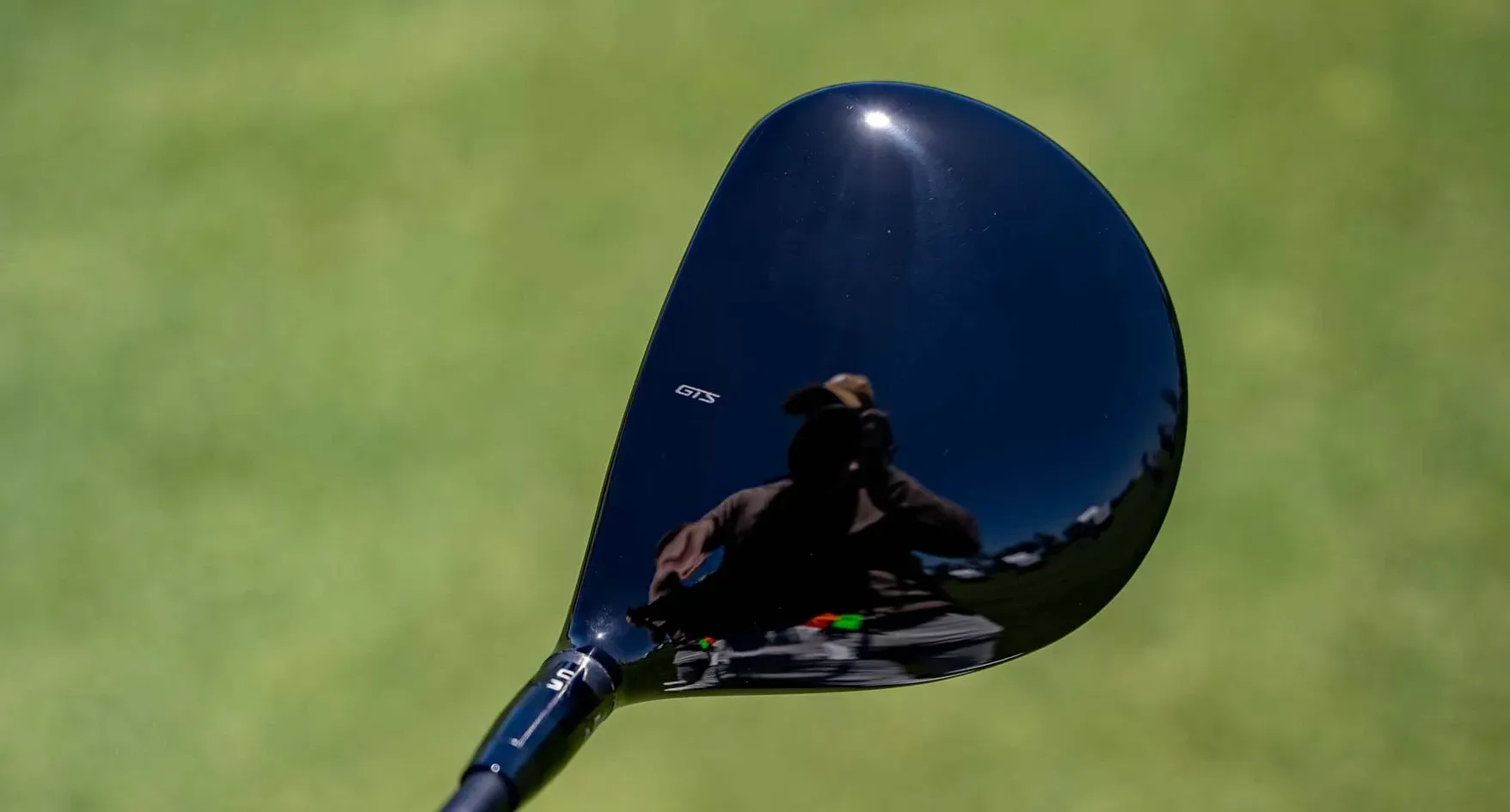 Close-up of a black Titleist GTS300 Mini Driver head reflecting a person with a hat holding a camera, outdoors on a green grassy field.