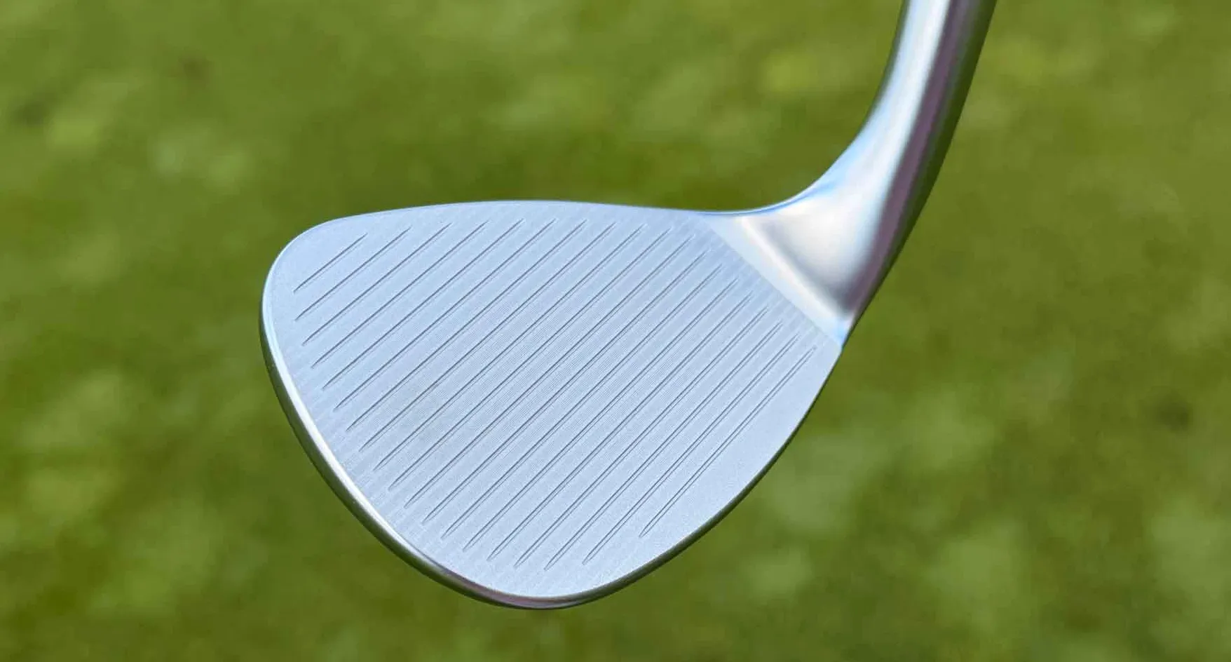 Close-up of a clean, silver Callaway Full Toe golf club wedge head with visible grooves, set against a blurred green grass background.