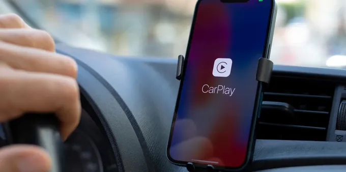 iPhone mounted on a car dashboard displaying the Apple CarPlay connection screen while a driver holds the steering wheel.