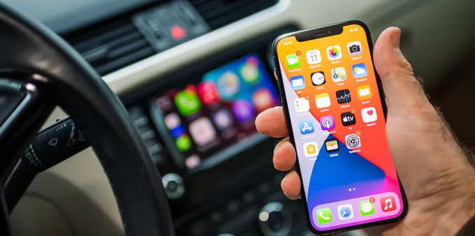 A hand holding an iPhone with a blurred CarPlay display in the background.