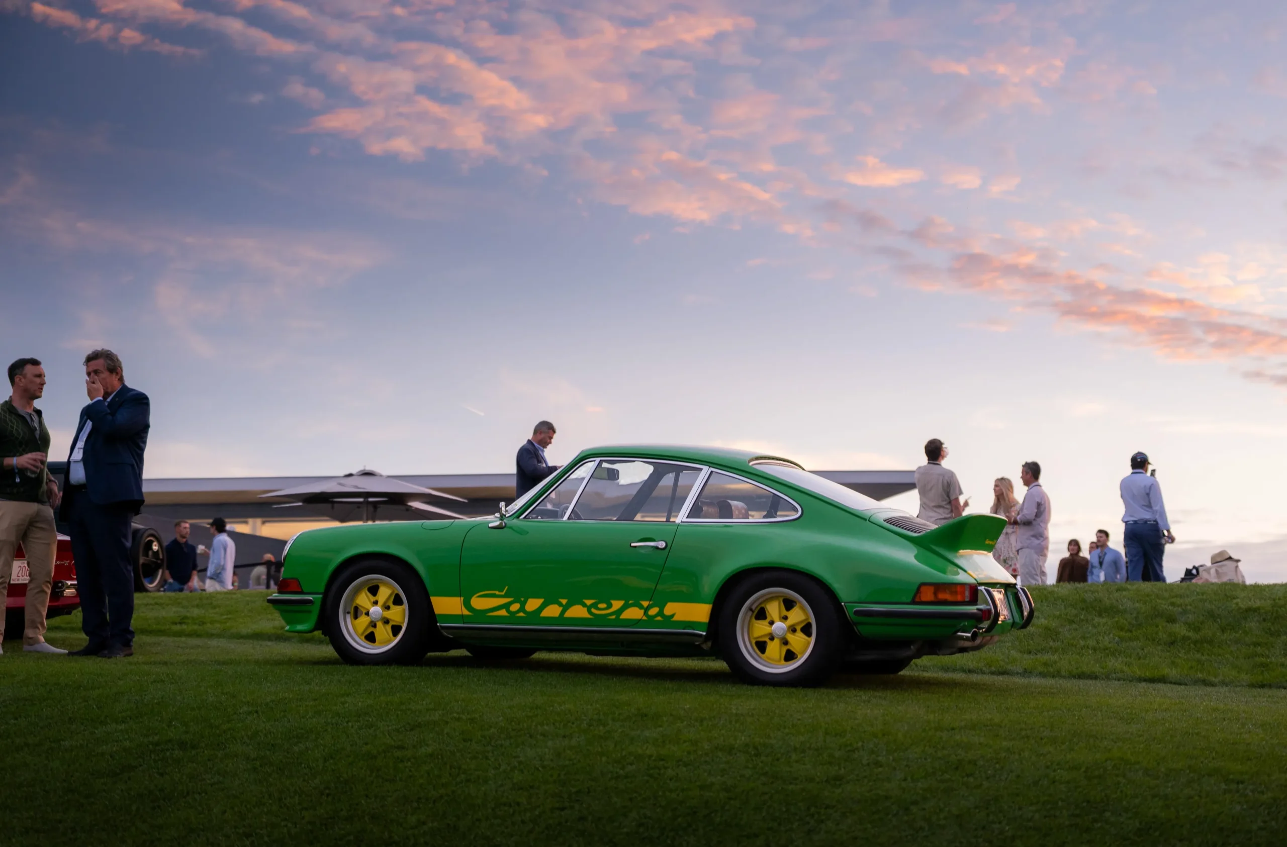 A bright green vintage car with yellow accents parked on grass, with people gathered in the background under a sunset sky.