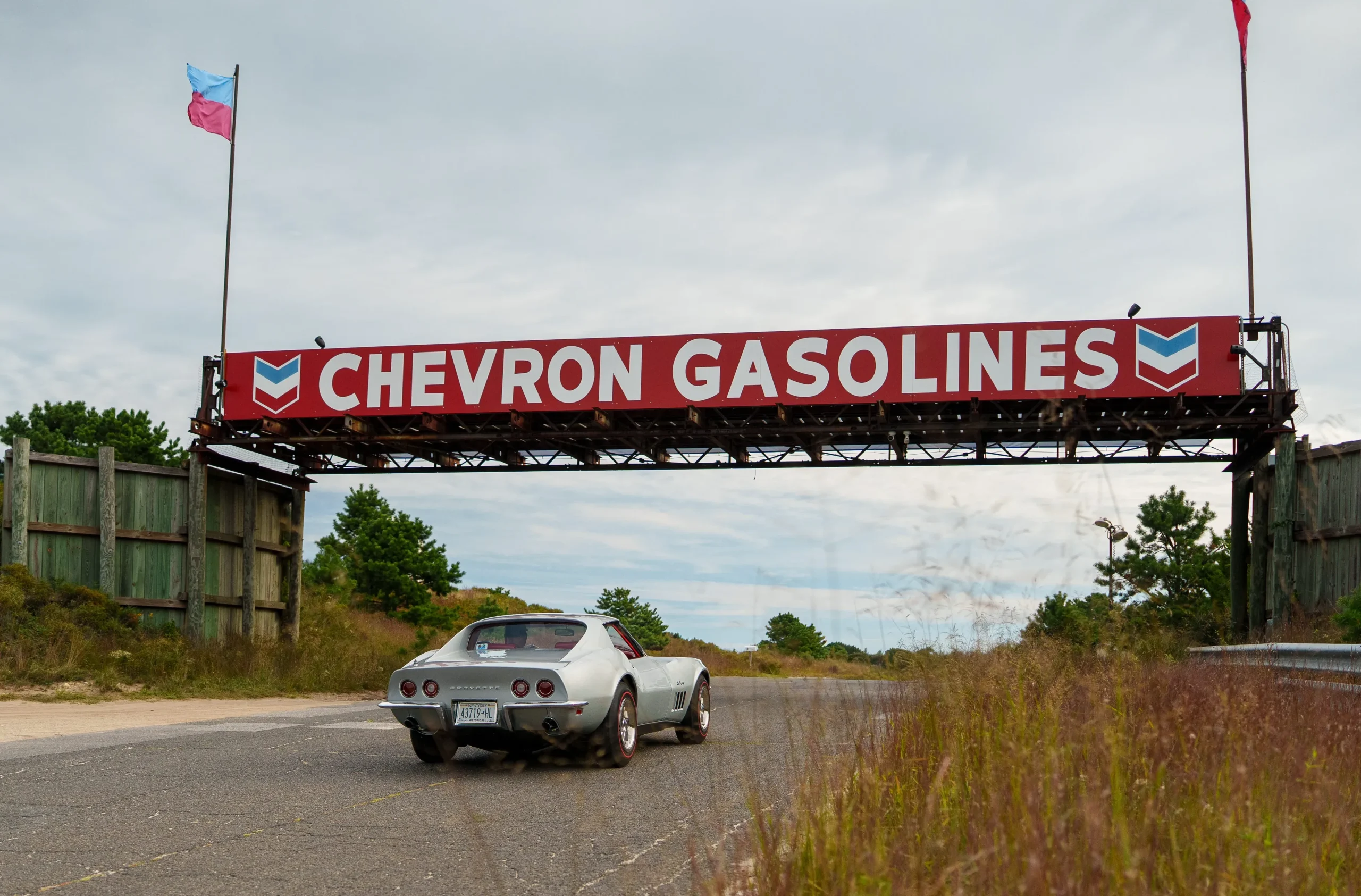 A classic car parked beneath a Chevron Gasolines sign along a deserted road with greenery in the background.